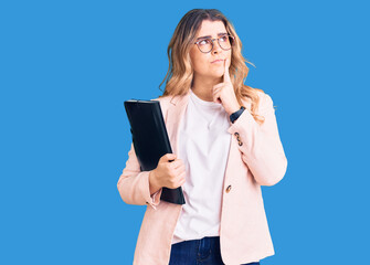 Young caucasian woman wearing business clothes and glasses holding binder serious face thinking...