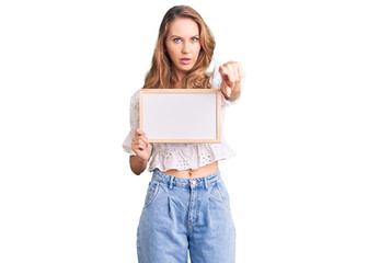 Fototapeta premium Young caucasian woman with blond hair holding blank empty banner pointing with finger to the camera and to you, confident gesture looking serious