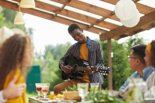 Portrait Of Young African-American Man Playing Guitar While Standing By Table And Enjoying Dinner With Friends Outdoors At Summer Party, Copy Space