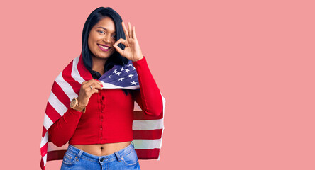 Hispanic woman with long hair holding united states flag doing ok sign with fingers, smiling...