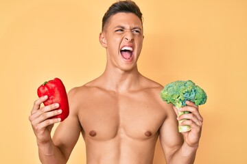 Young hispanic boy shirtless holding broccoli and red pepper angry and mad screaming frustrated and furious, shouting with anger. rage and aggressive concept.