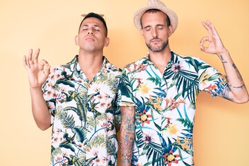 Young gay couple of two men wearing summer hat and hawaiian shirt relaxed and smiling with eyes closed doing meditation gesture with fingers. yoga concept.