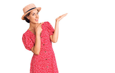 Young beautiful woman wearing summer hat amazed and smiling to the camera while presenting with hand and pointing with finger.