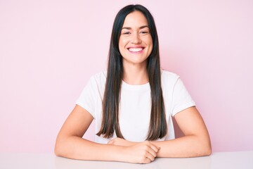 Fototapeta premium Young caucasian woman sitting at the table wearing casual white tshirt looking positive and happy standing and smiling with a confident smile showing teeth