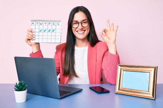 Young caucasian woman sitting at the desk holding travel calendar doing ok sign with fingers, smiling friendly gesturing excellent symbol