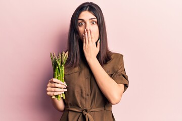 Young beautiful girl holding asparagus covering mouth with hand, shocked and afraid for mistake. surprised expression