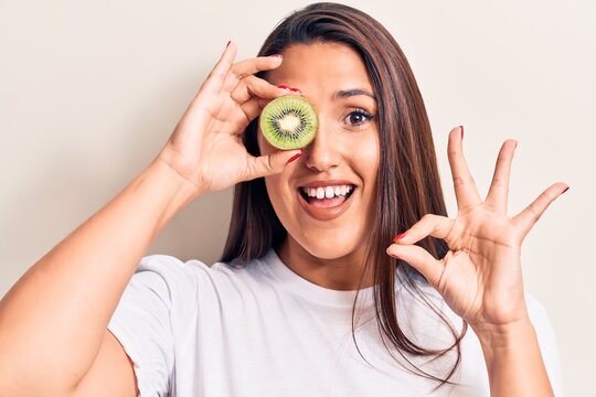 Young beautiful brunette woman holding kiwi doing ok sign with fingers, smiling friendly gesturing excellent symbol