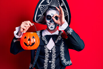Young man wearing mexican day of the dead costume holding pumpkin smiling happy doing ok sign with hand on eye looking through fingers