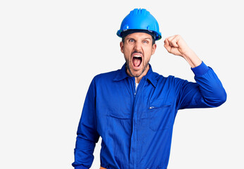 Young handsome man wearing worker uniform and hardhat crazy and mad shouting and yelling with aggressive expression and arms raised. frustration concept.