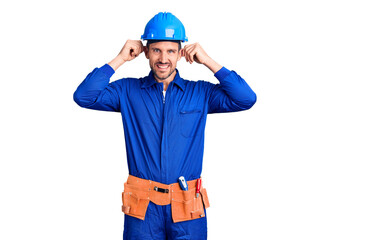 Young handsome man wearing worker uniform and hardhat smiling pulling ears with fingers, funny...