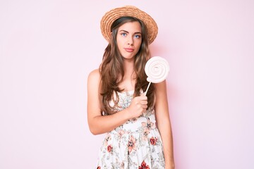 Beautiful young caucasian woman wearing summer hat and lollipop thinking attitude and sober...