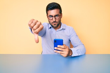 Young hispanic man using smartphone sitting on the table with angry face, negative sign showing dislike with thumbs down, rejection concept