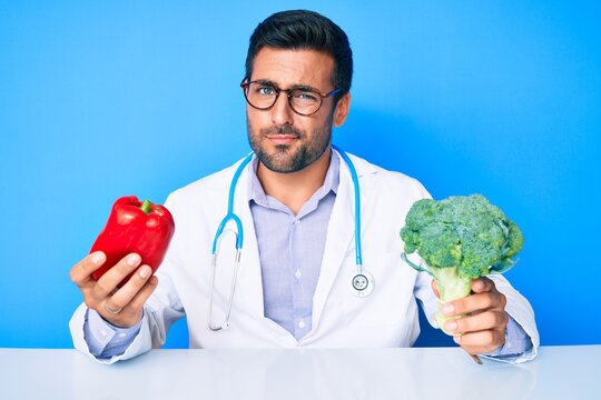 Young hispanic man wearing doctor uniform holding red pepper and broccoli skeptic and nervous, frowning upset because of problem. negative person.