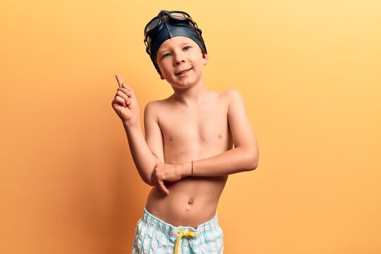 Cute blond kid wearing swimwear and swimmer glasses smiling happy pointing with hand and finger to the side