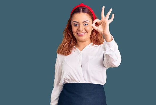 Young latin woman wearing waitress apron smiling positive doing ok sign with hand and fingers. successful expression.