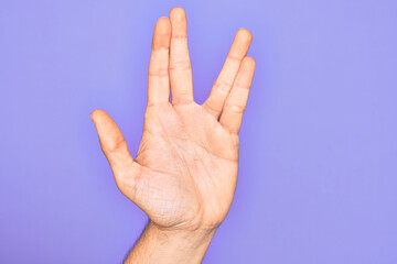 Hand of caucasian young man showing fingers over isolated purple background greeting doing Vulcan salute, showing hand palm and fingers, freak culture