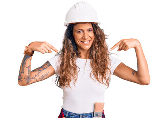 Young hispanic woman with tattoo wearing hardhat and builder clothes looking confident with smile...