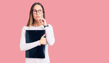 Beautiful brunette young woman holding business folder serious face thinking about question with...