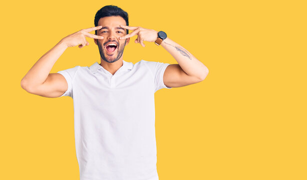 Young handsome hispanic man wearing casual clothes doing peace symbol with fingers over face, smiling cheerful showing victory