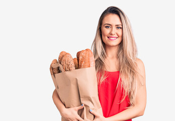 Young beautiful blonde woman holding paper bag with bread looking positive and happy standing and smiling with a confident smile showing teeth