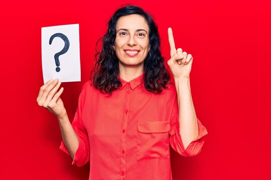 Young beautiful hispanic woman holding question mark smiling with an idea or question pointing finger with happy face, number one