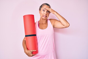 Middle age hispanic woman holding yoga mat stressed and frustrated with hand on head, surprised and angry face