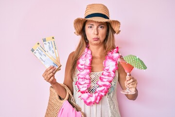 Middle age hispanic woman wearing hawaiian lei holding boarding pass and cocktail depressed and...
