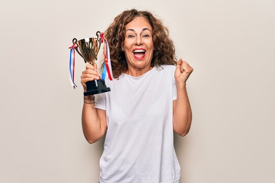 Middle age beautiful successful woman holding trophy for victory over white background screaming proud, celebrating victory and success very excited with raised arm