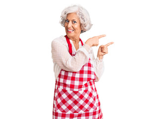 Senior grey-haired woman wearing apron smiling and looking at the camera pointing with two hands and fingers to the side.