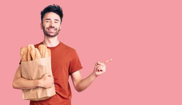 Young hispanic man holding paper bag with bread smiling happy pointing with hand and finger to the side