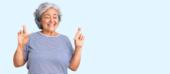 Senior woman with gray hair wearing casual striped clothes gesturing finger crossed smiling with hope and eyes closed. luck and superstitious concept.