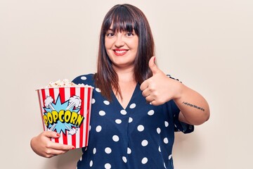 Young plus size woman holding popcorn smiling happy and positive, thumb up doing excellent and approval sign