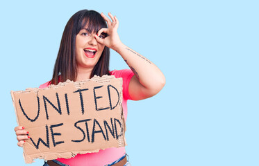 Young plus size woman holding united we stand banner smiling happy doing ok sign with hand on eye looking through fingers