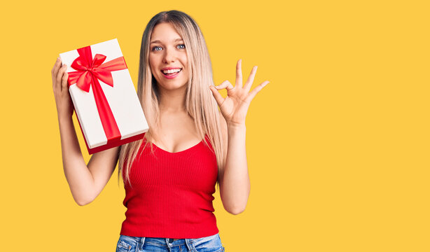 Young beautiful blonde woman holding gift doing ok sign with fingers, smiling friendly gesturing excellent symbol