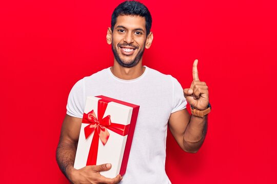 Young latin man holding gift smiling with an idea or question pointing finger with happy face, number one
