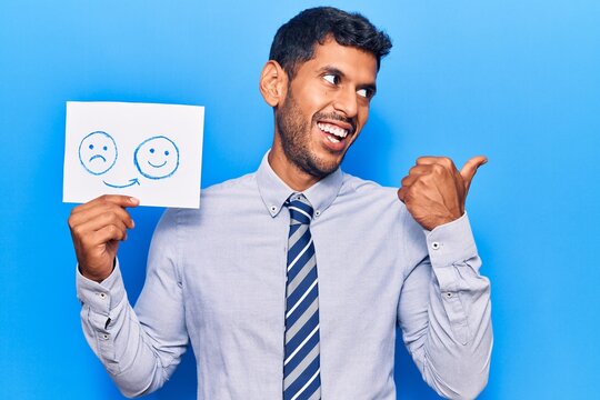 Young latin man holding sad to happy emotion paper pointing thumb up to the side smiling happy with open mouth