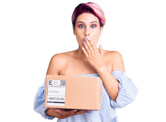 Young beautiful woman with pink hair holding delivery package covering mouth with hand, shocked and...