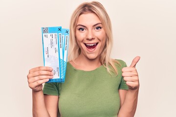 Beautiful blonde tourist woman on vacation holding airline boarding pass over white background smiling happy and positive, thumb up doing excellent and approval sign