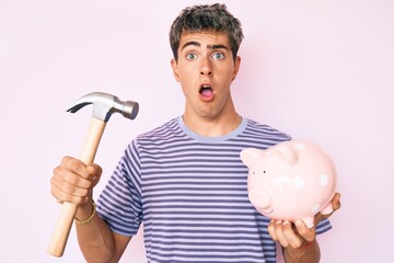 Young handsome man holding piggy bank and hammer afraid and shocked with surprise and amazed expression, fear and excited face.