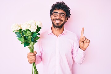 Handsome young man with curly hair and bear holding bouquet of white flowers smiling with an idea or question pointing finger with happy face, number one