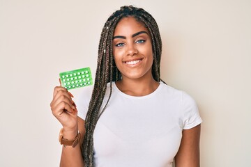Young african american woman with braids holding birth control pills looking positive and happy...