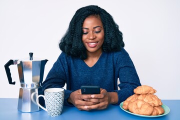Beautiful african woman eating breakfast using smartphone looking positive and happy standing and smiling with a confident smile showing teeth