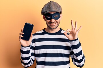 Young handsome bald man wearing burglar mask holding smartphone doing ok sign with fingers, smiling friendly gesturing excellent symbol