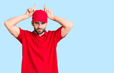 Naklejka premium Young handsome man with beard wearing delivery uniform doing funny gesture with finger over head as bull horns