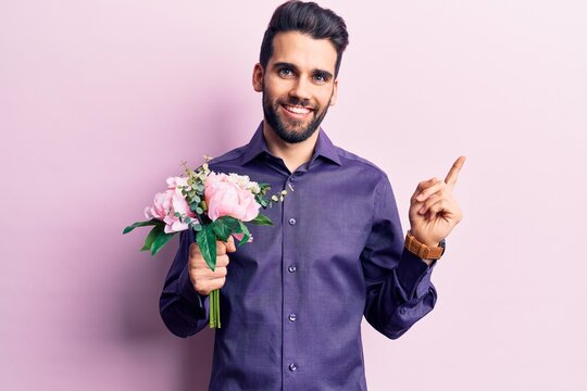 Young handsome man with beard holding bouquet of flowers smiling happy pointing with hand and finger to the side