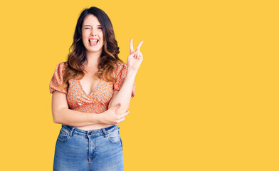 Beautiful young brunette woman wearing casual clothes smiling with happy face winking at the camera doing victory sign. number two.