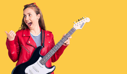 Fototapeta premium Young beautiful blonde woman playing electric guitar pointing thumb up to the side smiling happy with open mouth
