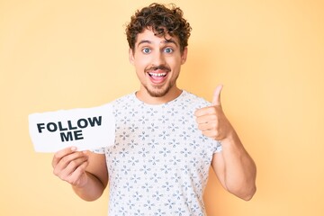 Young caucasian man with curly hair holding follow me message paper smiling happy and positive, thumb up doing excellent and approval sign