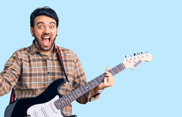 Young hispanic man playing electric guitar screaming proud, celebrating victory and success very excited with raised arms