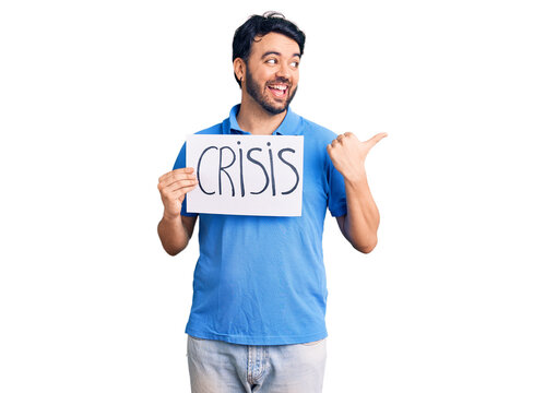 Young hispanic man holding crisis message paper pointing thumb up to the side smiling happy with open mouth